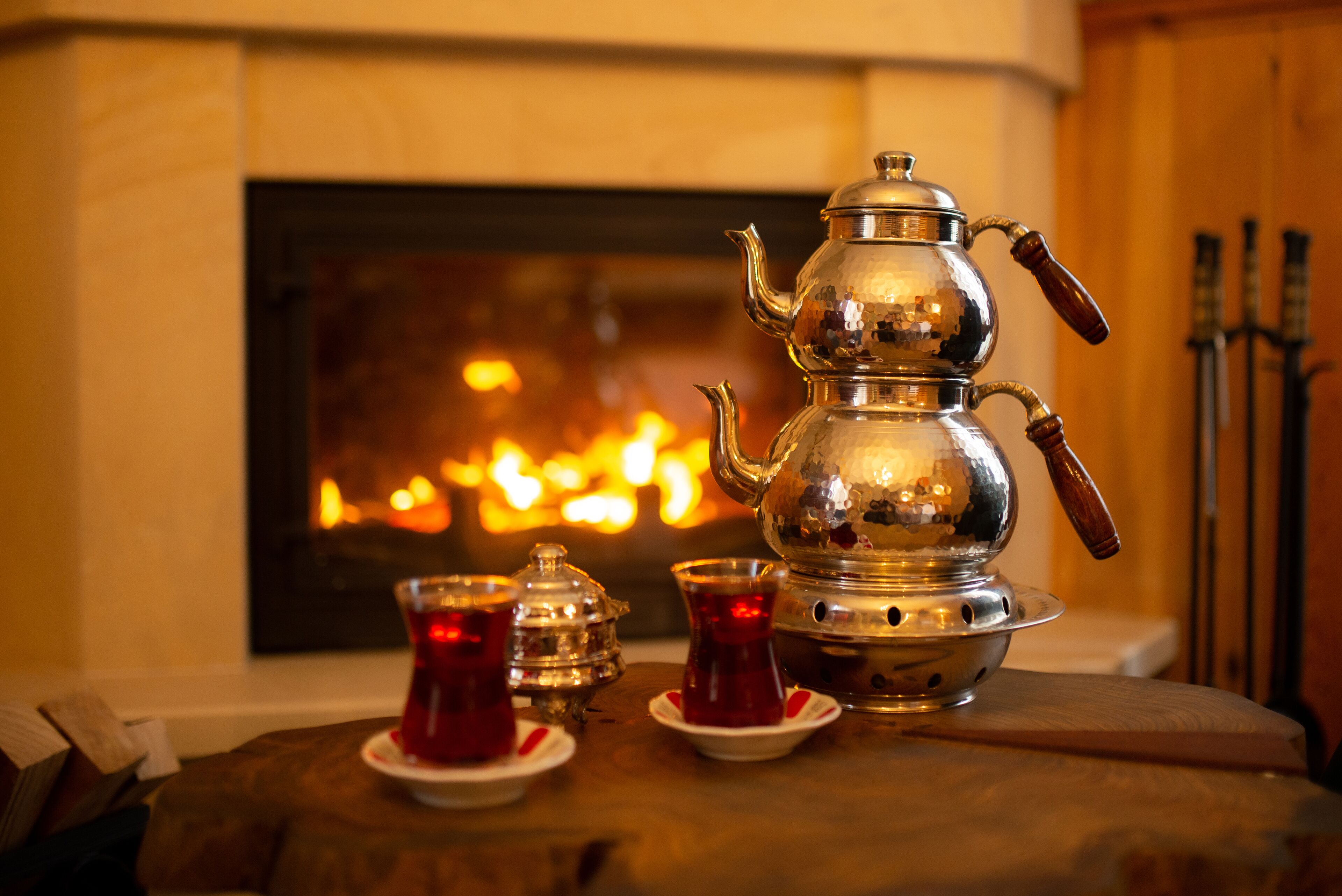 A traditional metal teapot with a wooden handle, accompanied by two glass cups with red liquid, set on a wooden table in front of a fireplace.