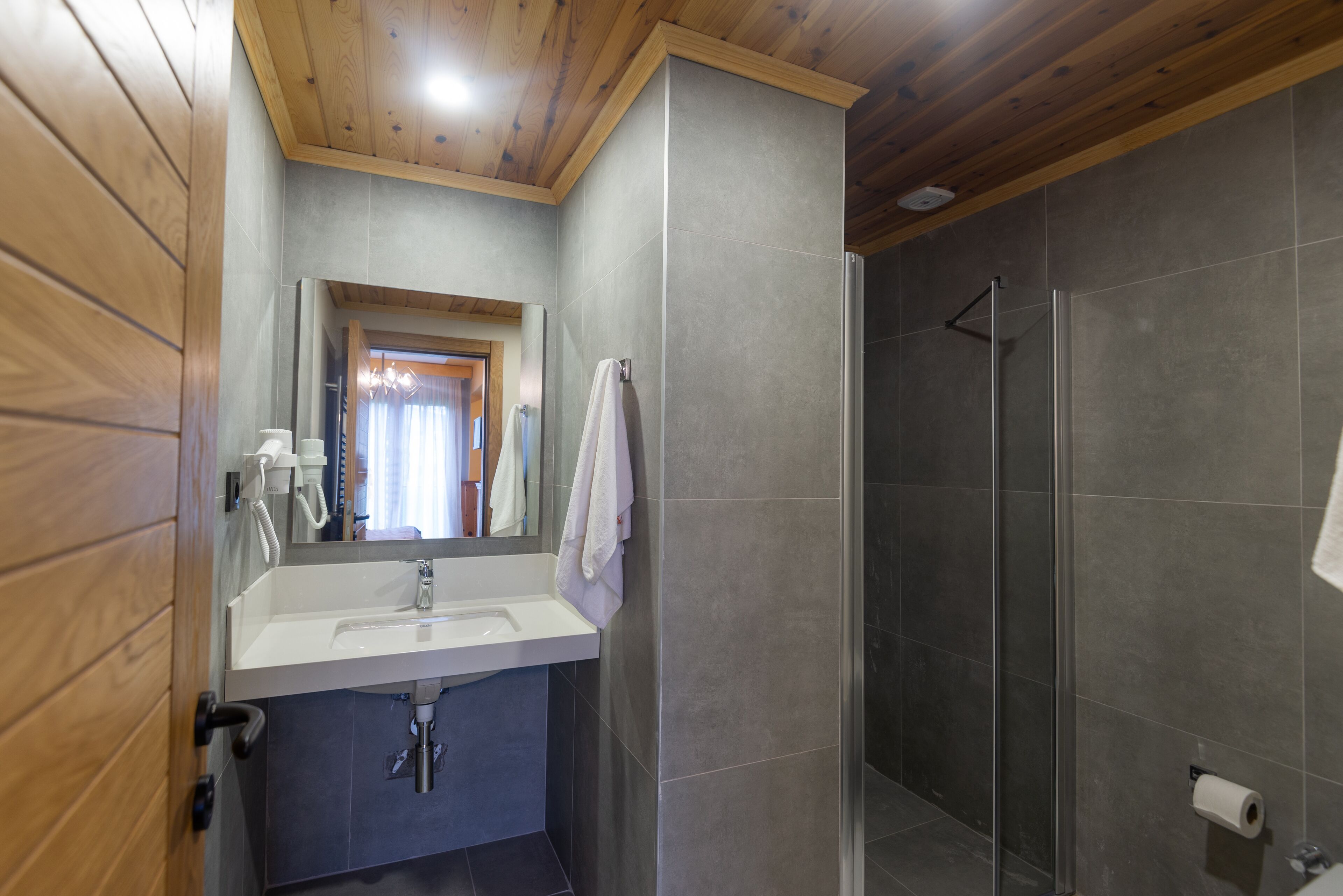 A bathroom with a wooden ceiling, a sink, a mirror, and a shower area.