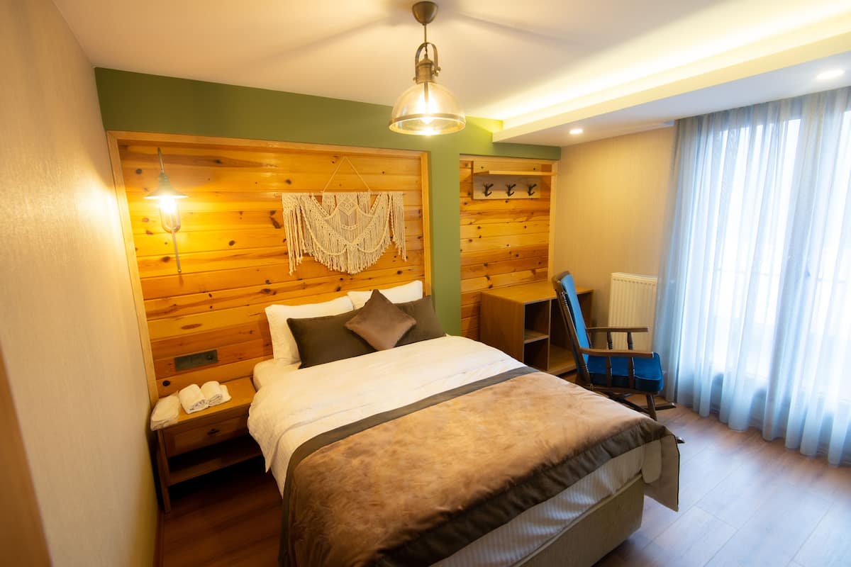 A hotel room with a wooden headboard, a bed with a brown blanket, a nightstand with towels, a wooden chair, and a window with sheer curtains.