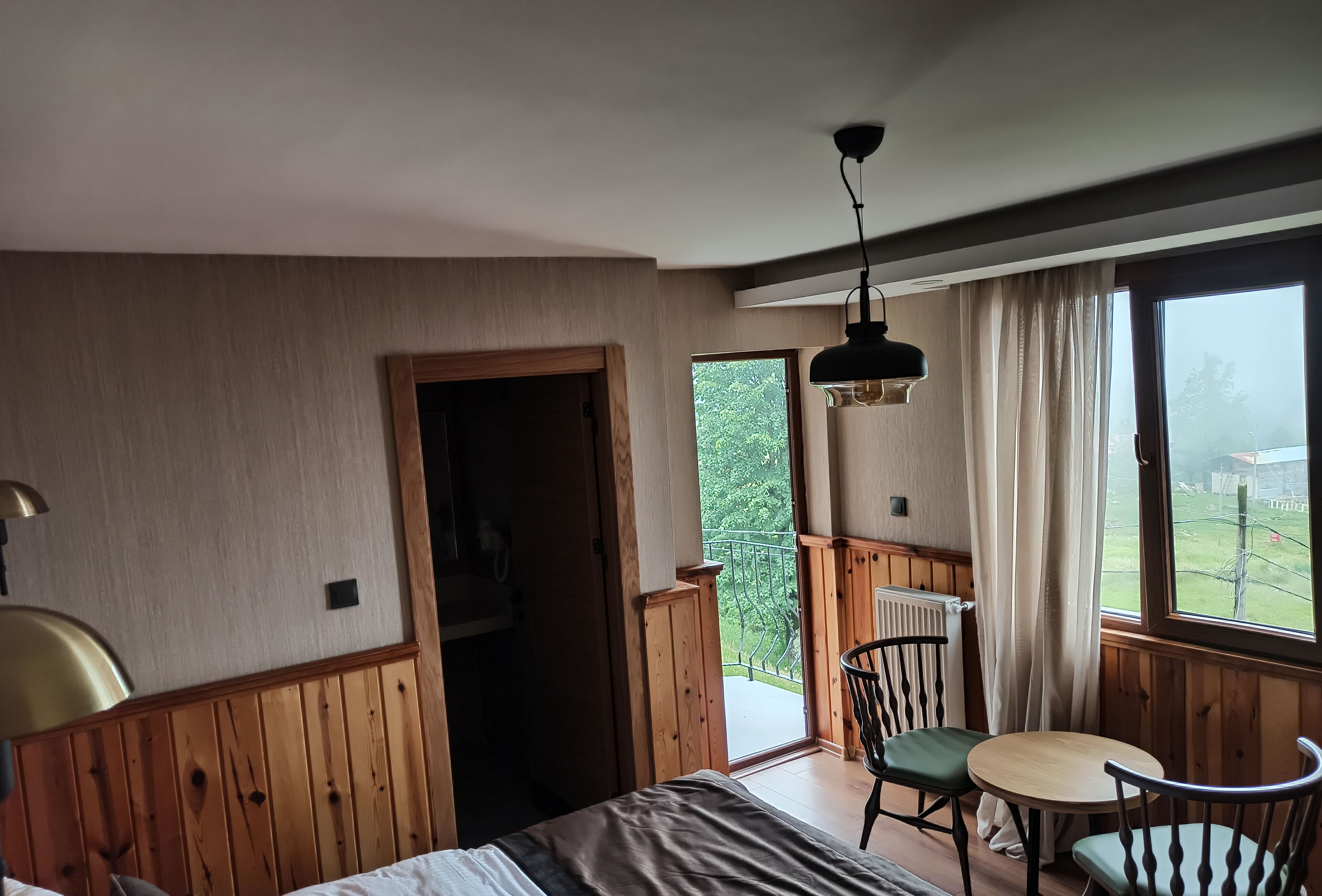 A room with a bed, a table, two chairs, a hanging light fixture, and a view of a garden through a window.