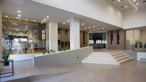 Un lobby amplio con diseño moderno, que incluye una escalera central, un área de descanso con sillas amarillas y una cafetería visible a través del muro de cristal.