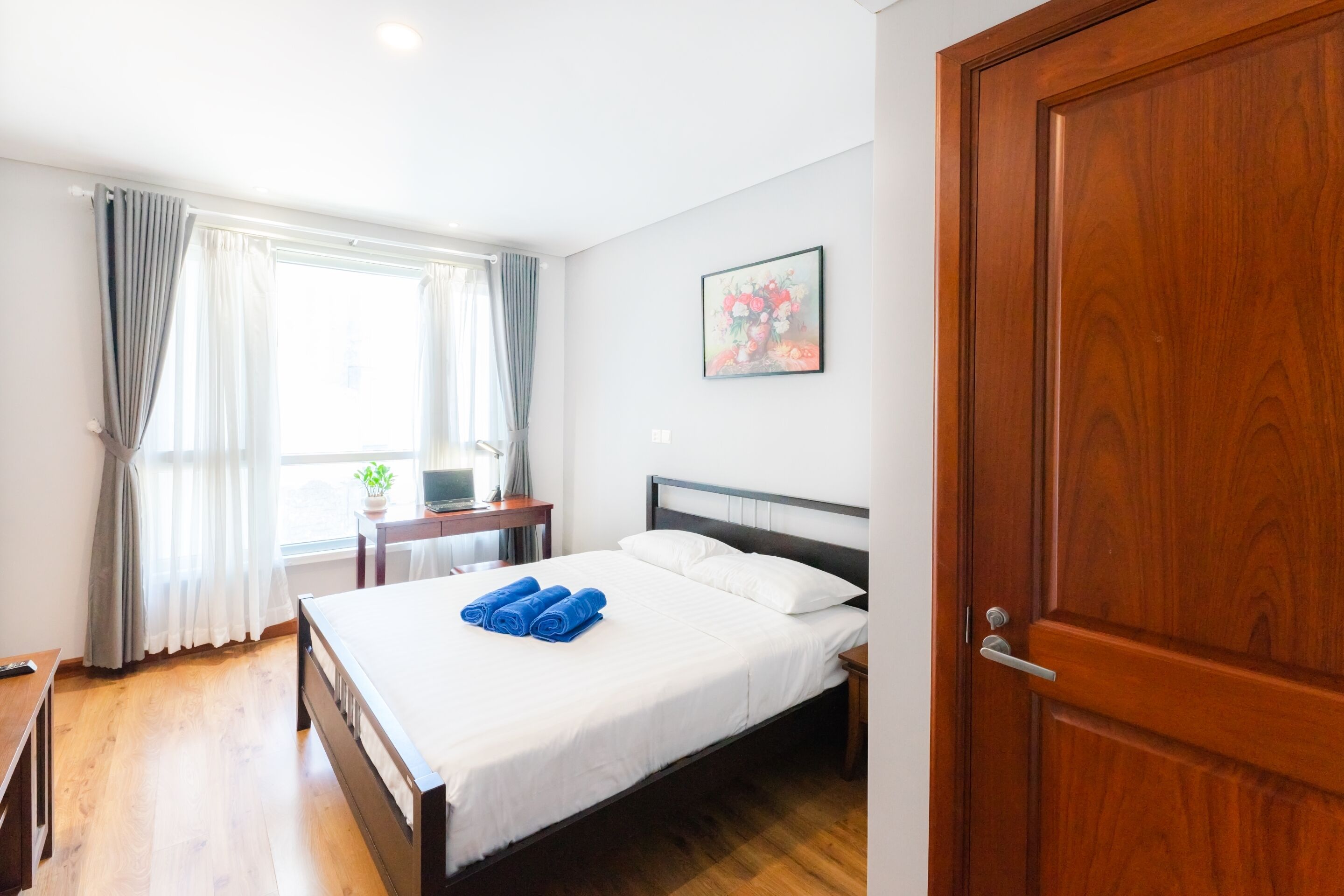 A hotel room with a wooden bed, a desk with a computer, a window with curtains, and a door.