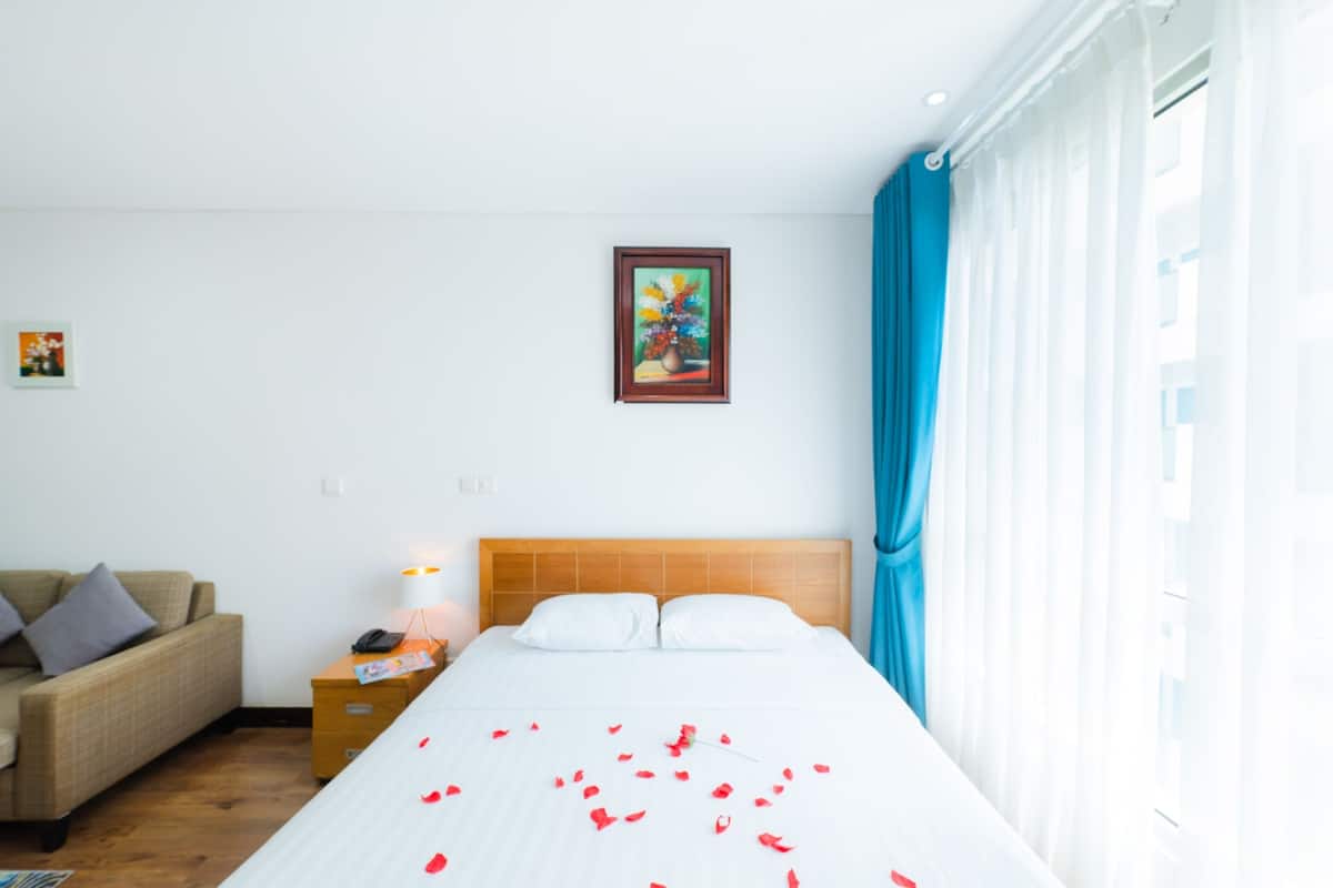 A hotel room with a bed, a nightstand, a lamp, a painting on the wall, and a window with curtains.