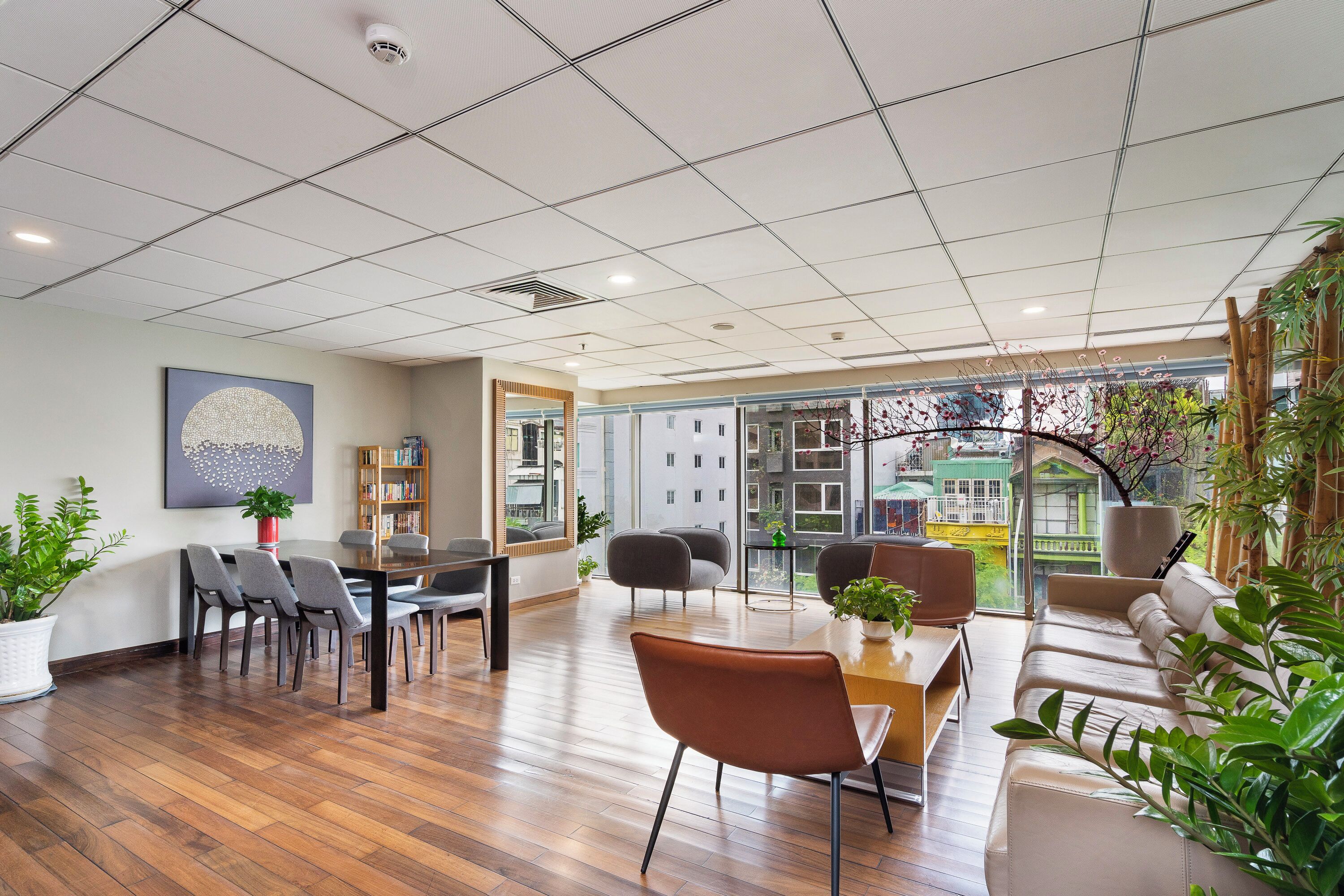 A spacious office with a wooden floor, a large table with chairs, a sofa, and a bookshelf. There are potted plants and a large window with a view of buildings.