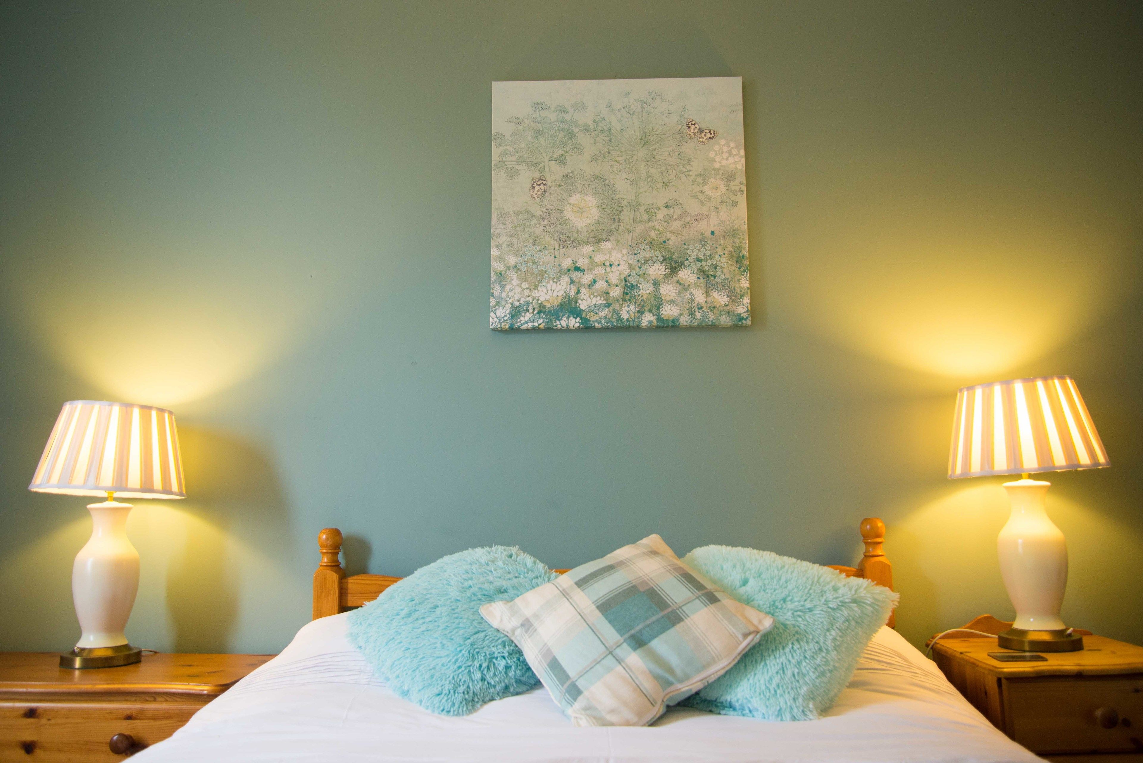 A bedroom with a bed, two bedside lamps, a floral artwork on the wall, and a blue fluffy pillow.