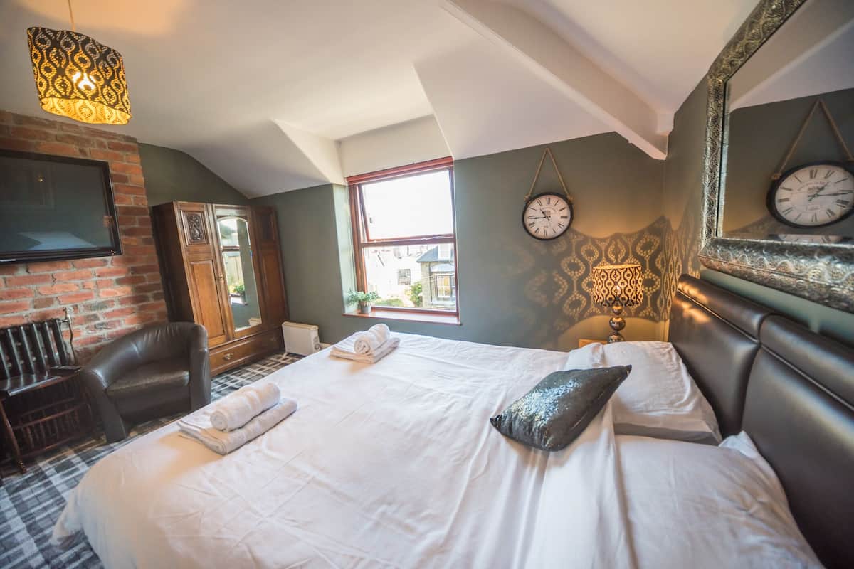 A neatly made bed with white linens, a leather armchair, a large mirror with clocks, and a patterned wall lamp.