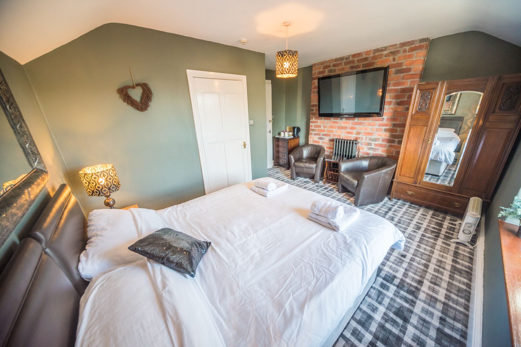 A bedroom with a large bed, a TV mounted on a brick wall, a leather armchair, and a mirror.