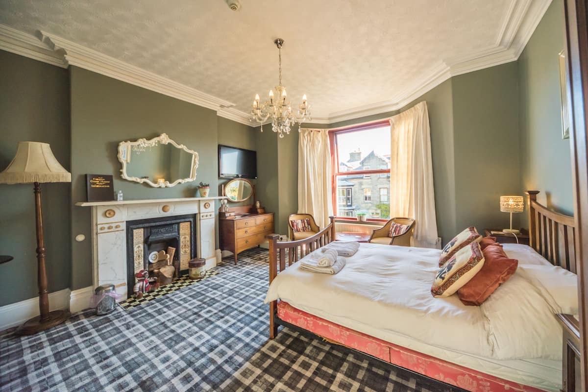 A hotel room with a checkered floor, a bed with pillows, a dresser, a mirror, a fireplace, and a view of buildings outside.