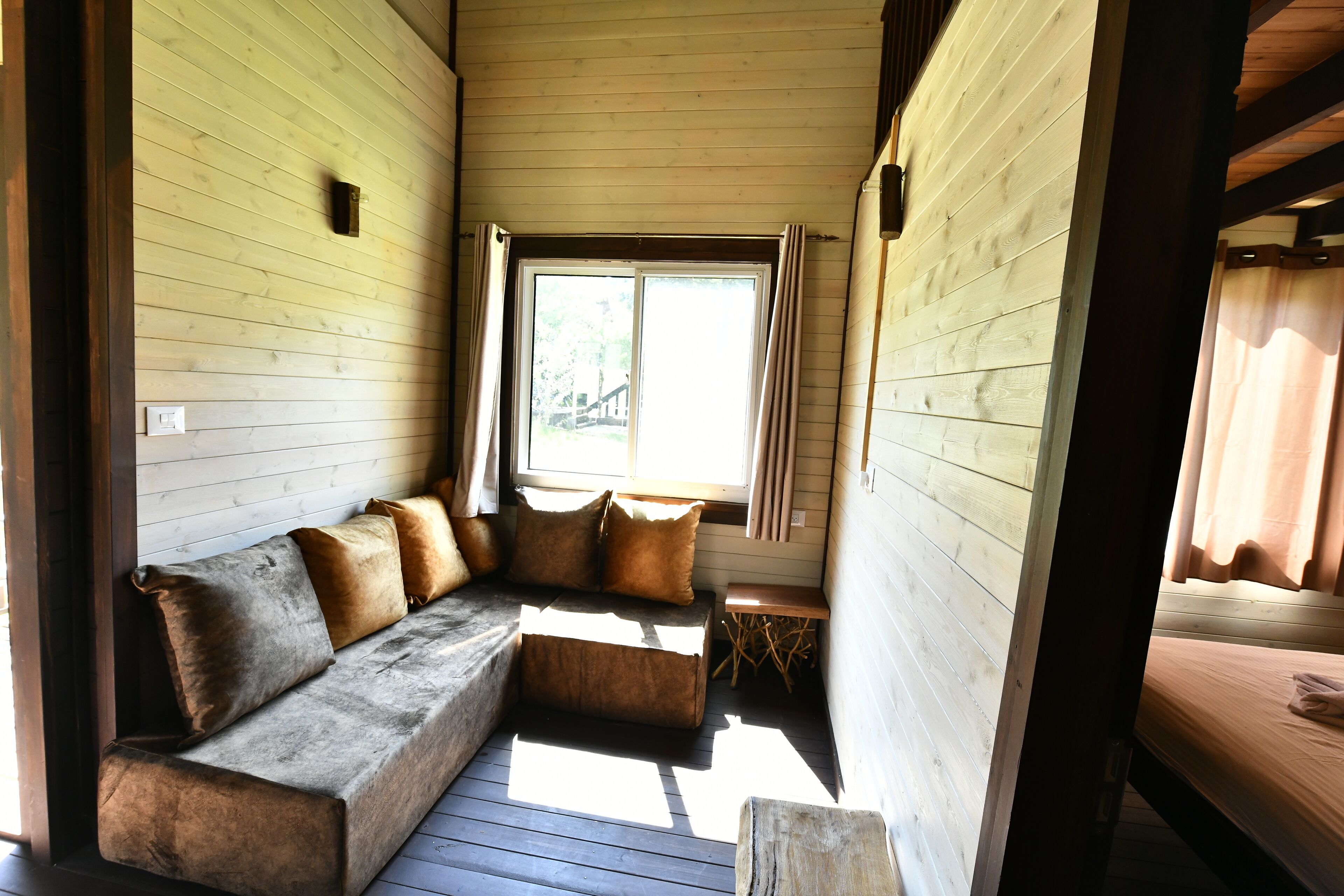 Interior acolhedor de uma cabana com soalho de madeira, um banco embutido com almofadas, uma pequena mesa e uma janela com cortinas.