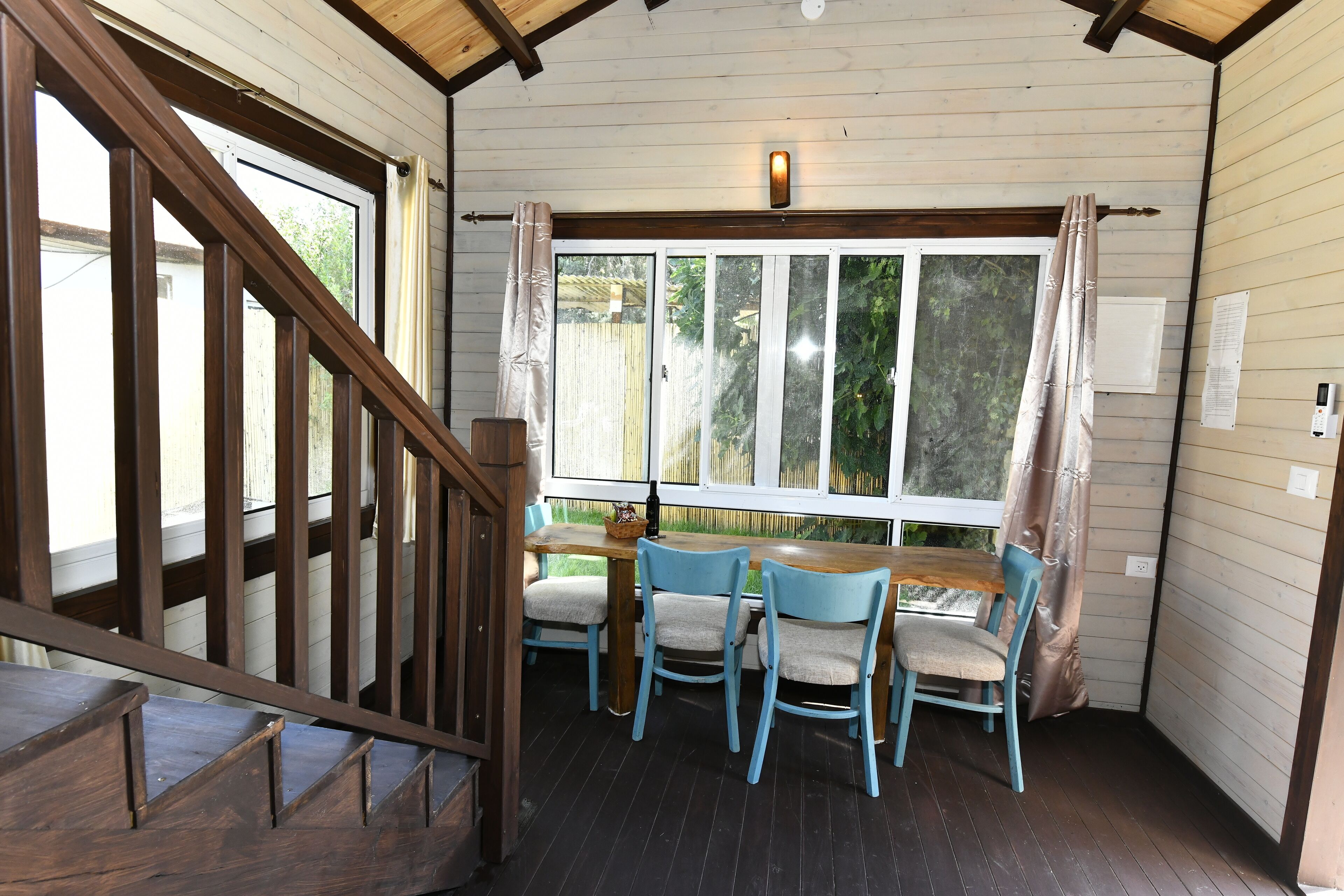 A wooden interior with a dining area featuring a table and chairs, a window with curtains, and a wooden staircase.