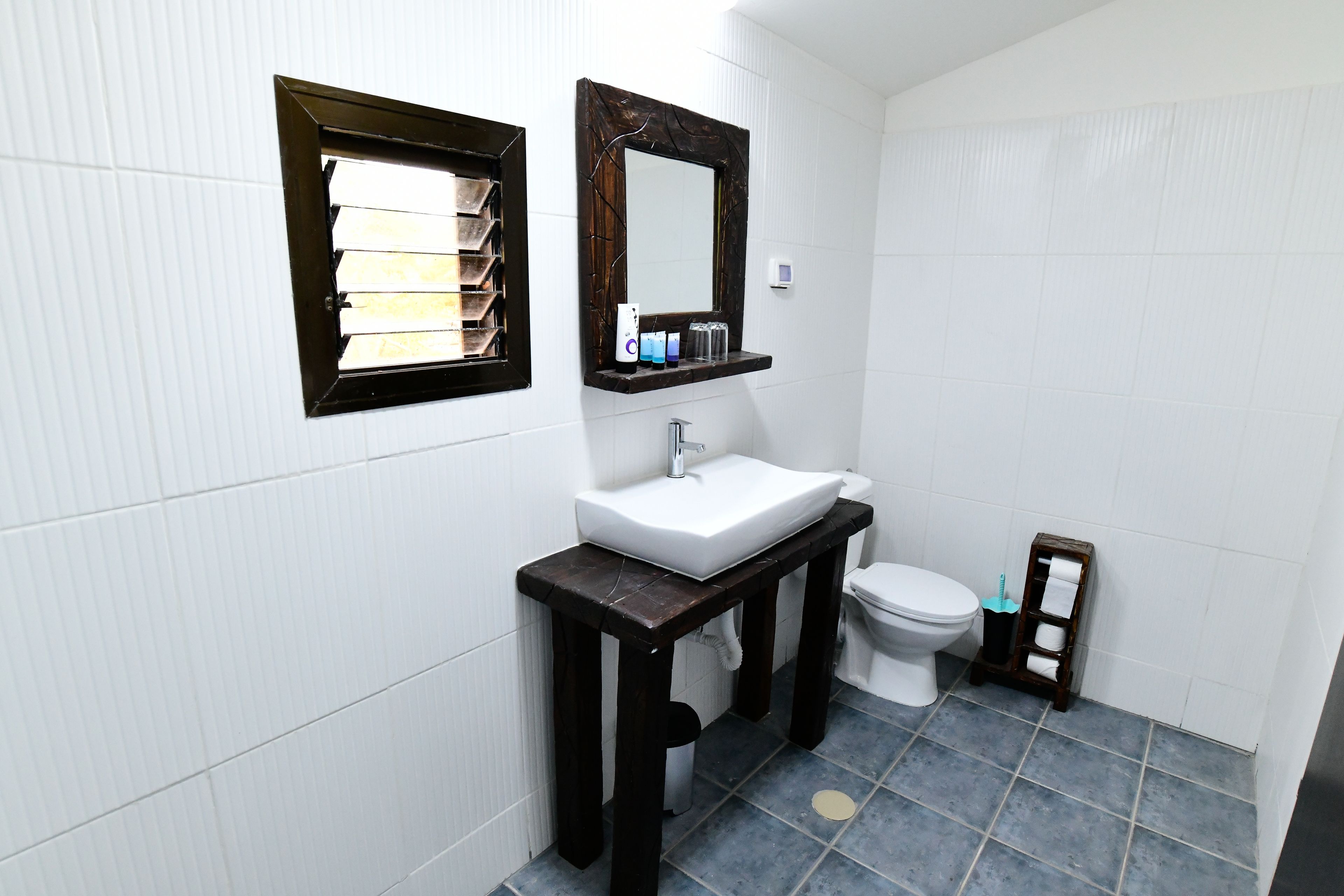 Et badeværelse med en hvid vask, et spejl, et toilet og en lille hylde med toiletartikler.