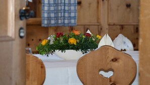 Uma mesa de madeira com toalha branca, um arranjo floral e recortes em formato de coração nas cadeiras.