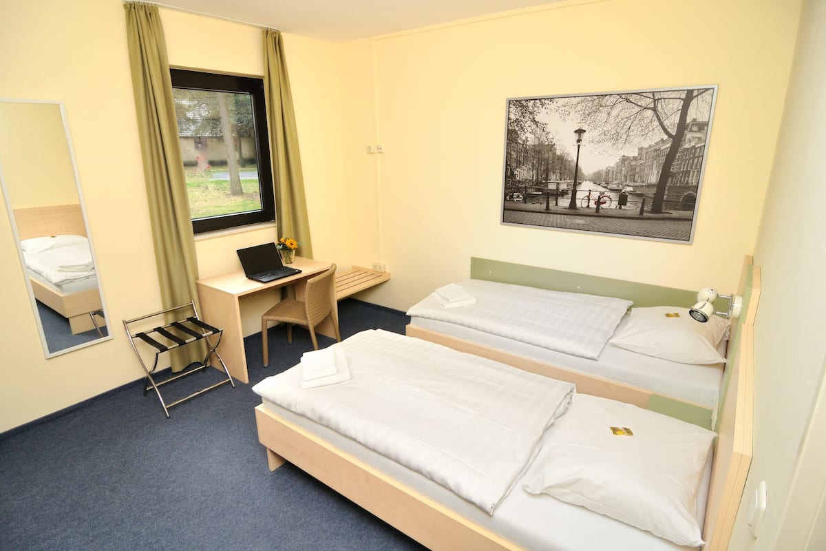 A hotel room with two beds, a desk with a laptop, a chair, a window with curtains, a mirror, and a wall-mounted lamp.