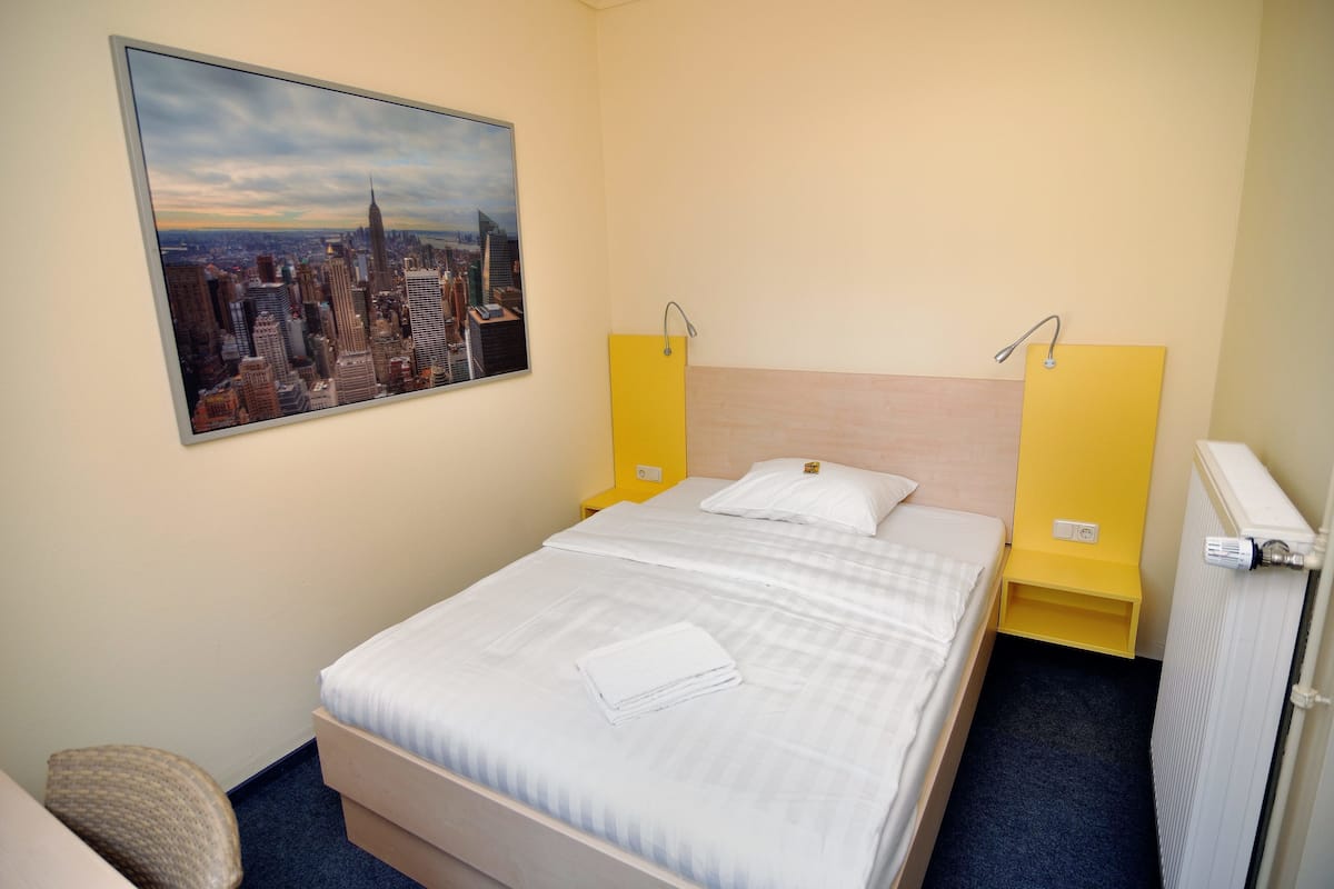 A hotel room with a bed, a cityscape painting, a yellow nightstand, and a radiator.