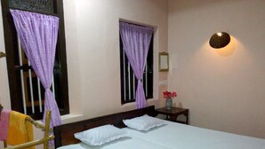 Un dormitorio con cama, dos ventanas con cortinas moradas, una mesita de noche con un florero y una lámpara fijada a la pared.