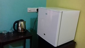 Un refrigerador blanco pequeño con un panel de control en la puerta, una tetera y dos vasos sobre una mesa oscura, frente a una pared verde azulado y amarilla.