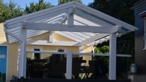 A covered patio area with a white roof structure, a wooden deck, and a yellow building in the background. - Ferienwohnung Bergisch Land (Wermelskirchen)