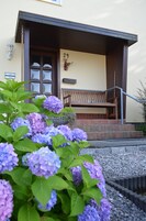 Um edifício com o número '29' na parede, um banco de madeira na parte externa e um canteiro de flores roxas.