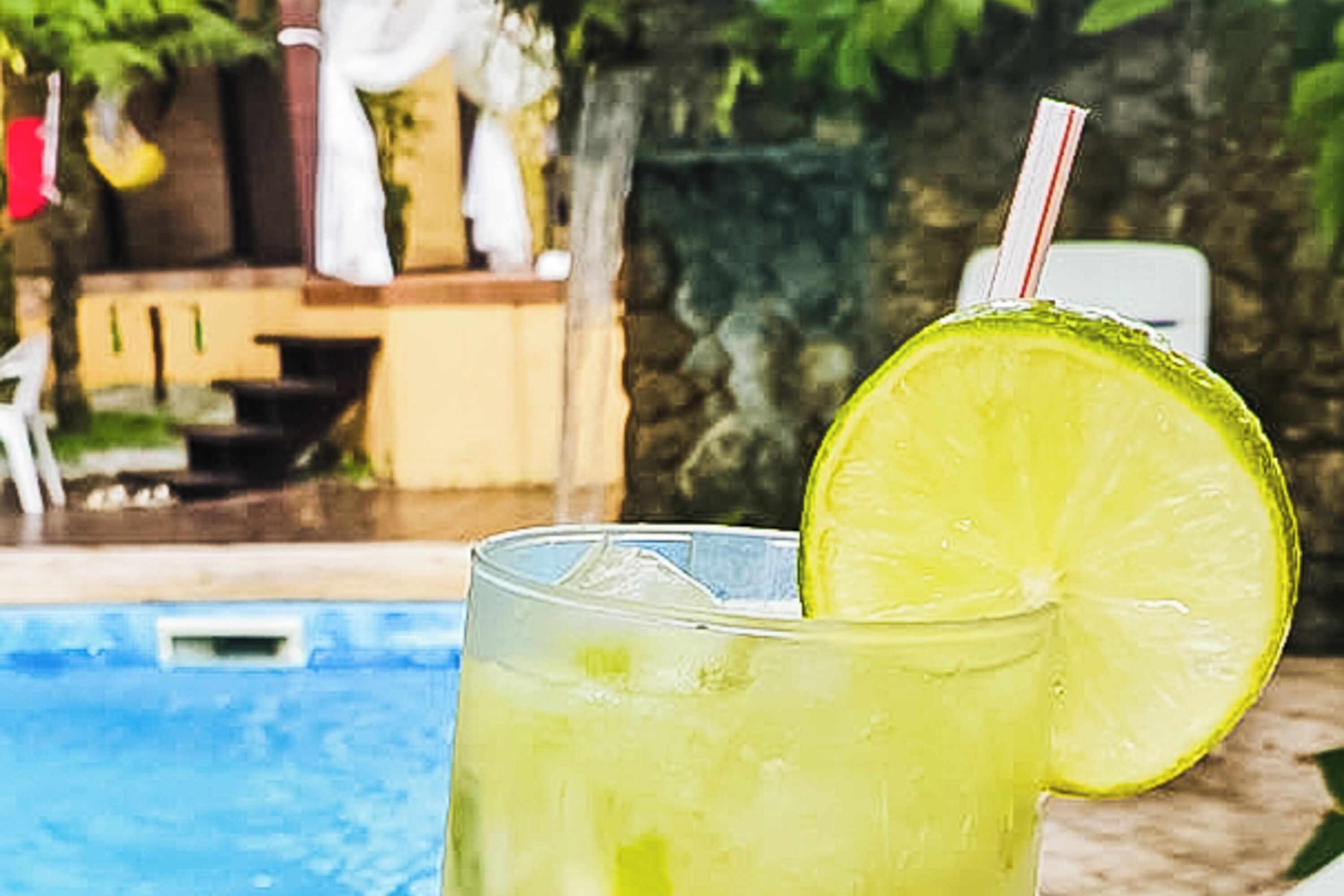 A glass of a yellowish beverage with a lime slice and a straw, placed on a poolside with a stone wall and a pool in the background.