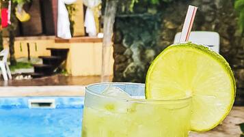 A glass of a yellowish beverage with a lime slice and a straw, placed on a poolside with a stone wall and a pool in the background.