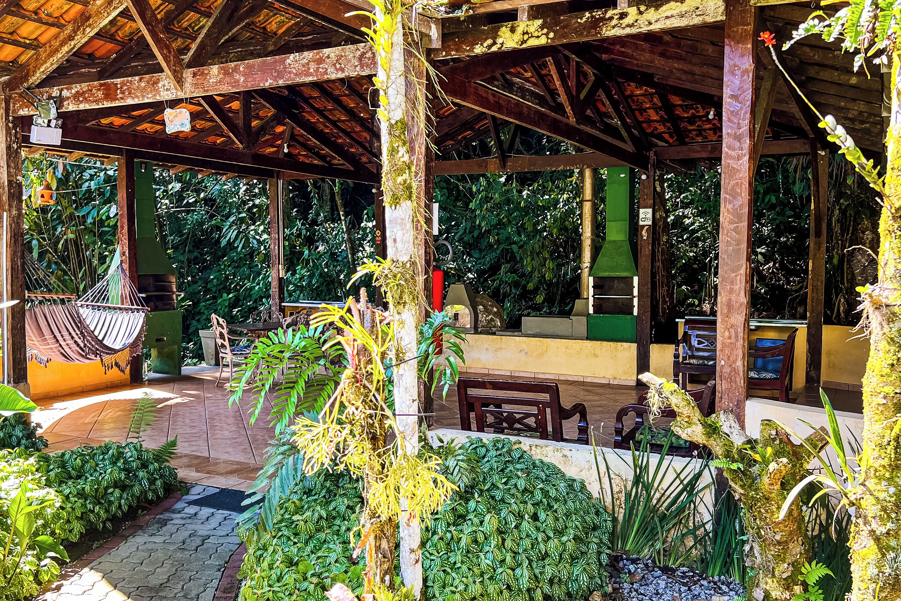 A covered outdoor area with wooden beams, a hammock, and various plants.
