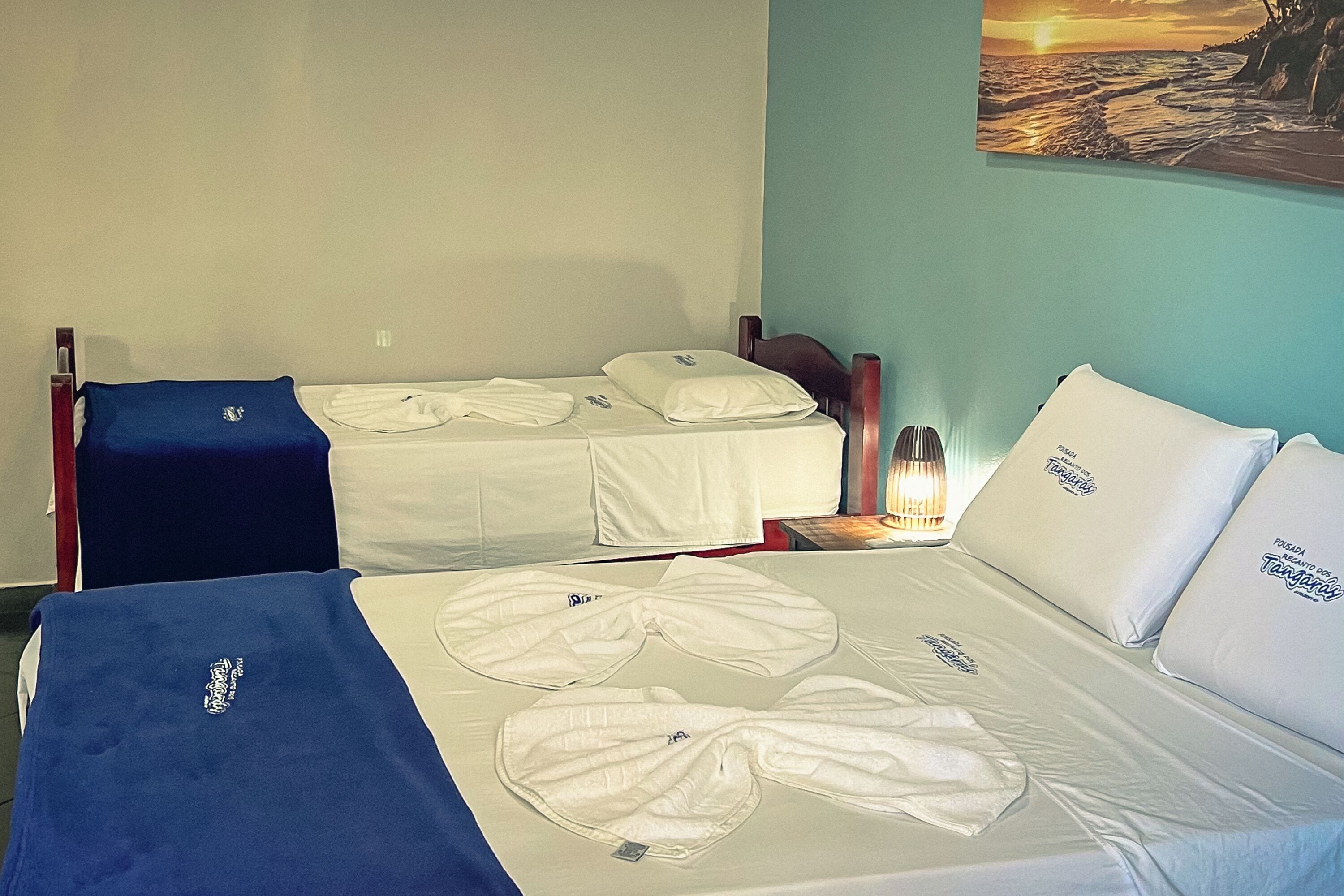 Quarto de hotel com duas camas de solteiro, cada uma com lençóis e almofadas brancas, um cobertor azul em uma cama e uma mesa de cabeceira com abajur.