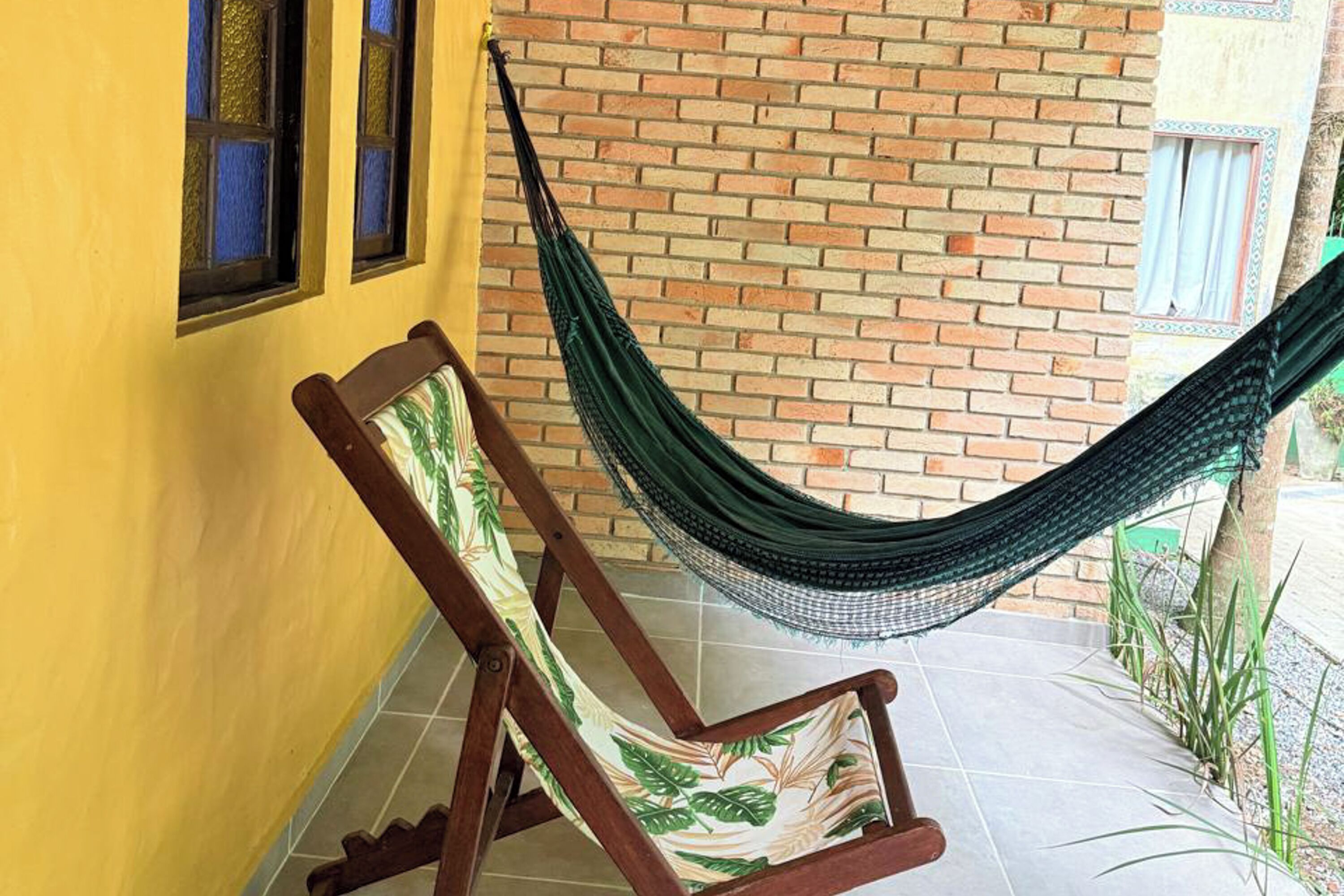 Una hamaca atada entre un muro de ladrillo y una silla de madera con un cojín estampado con motivos florales.