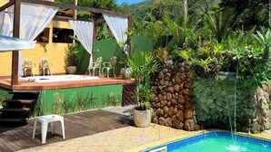 A pool area with a wooden deck, a small outdoor seating area with chairs, and a stone wall with a water feature. - VELINN Pousada Recanto dos Tangaras (São Sebastião)