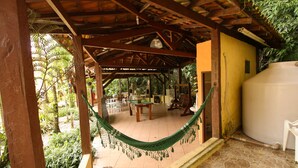A covered outdoor area with a hammock, a table, and chairs, surrounded by greenery and a yellow wall. - VELINN Pousada Recanto dos Tangaras (São Sebastião)