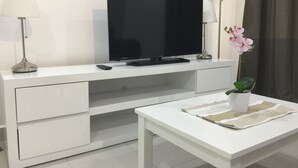 Un salon moderne avec un téléviseur à écran plat posé sur un meuble blanc, une télécommande, une lampe et une orchidée en pot sur une petite table.