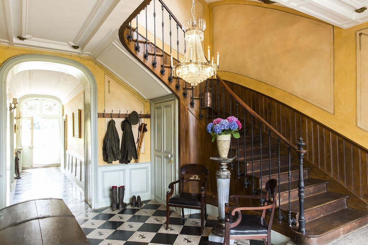A grand staircase with a chandelier, a checkered floor, and a coat rack with hats and boots.