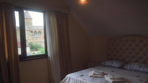 A hotel room with a bed, a window with curtains, a bedside table, and a small lamp. - Hotel Bagineti (Mtskheta)