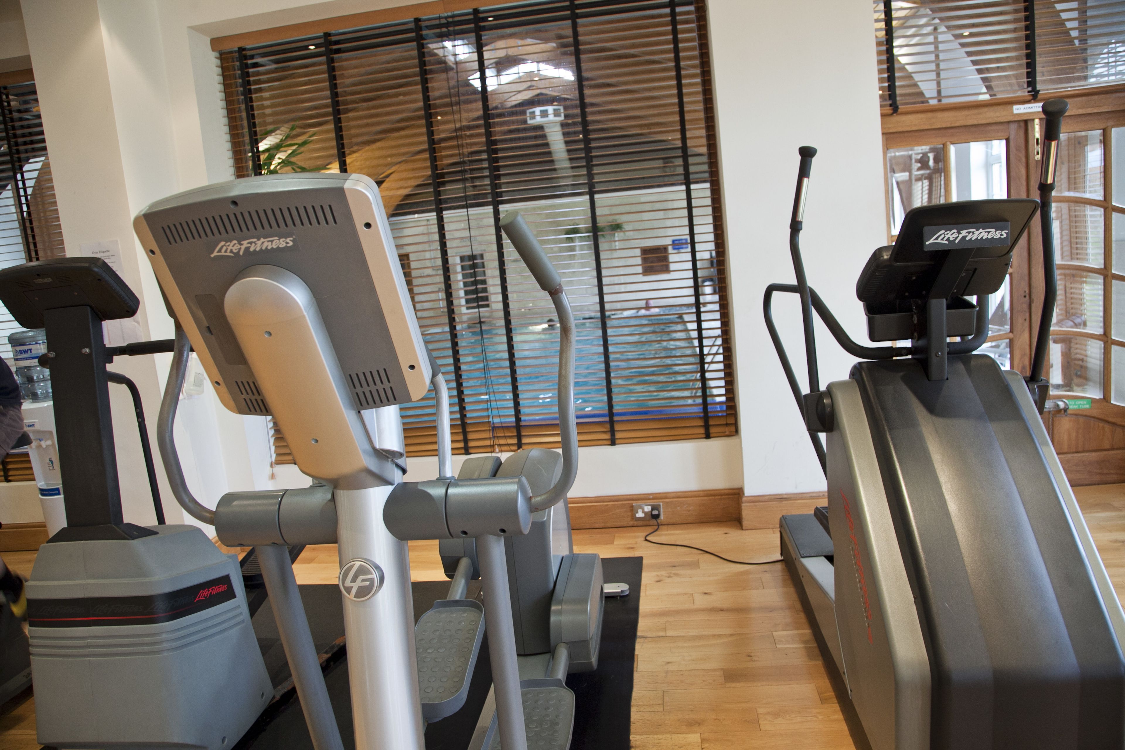 Ein Fitnessraum mit Cardiogeräten, Holzboden und einem Fenster mit Jalousien.
