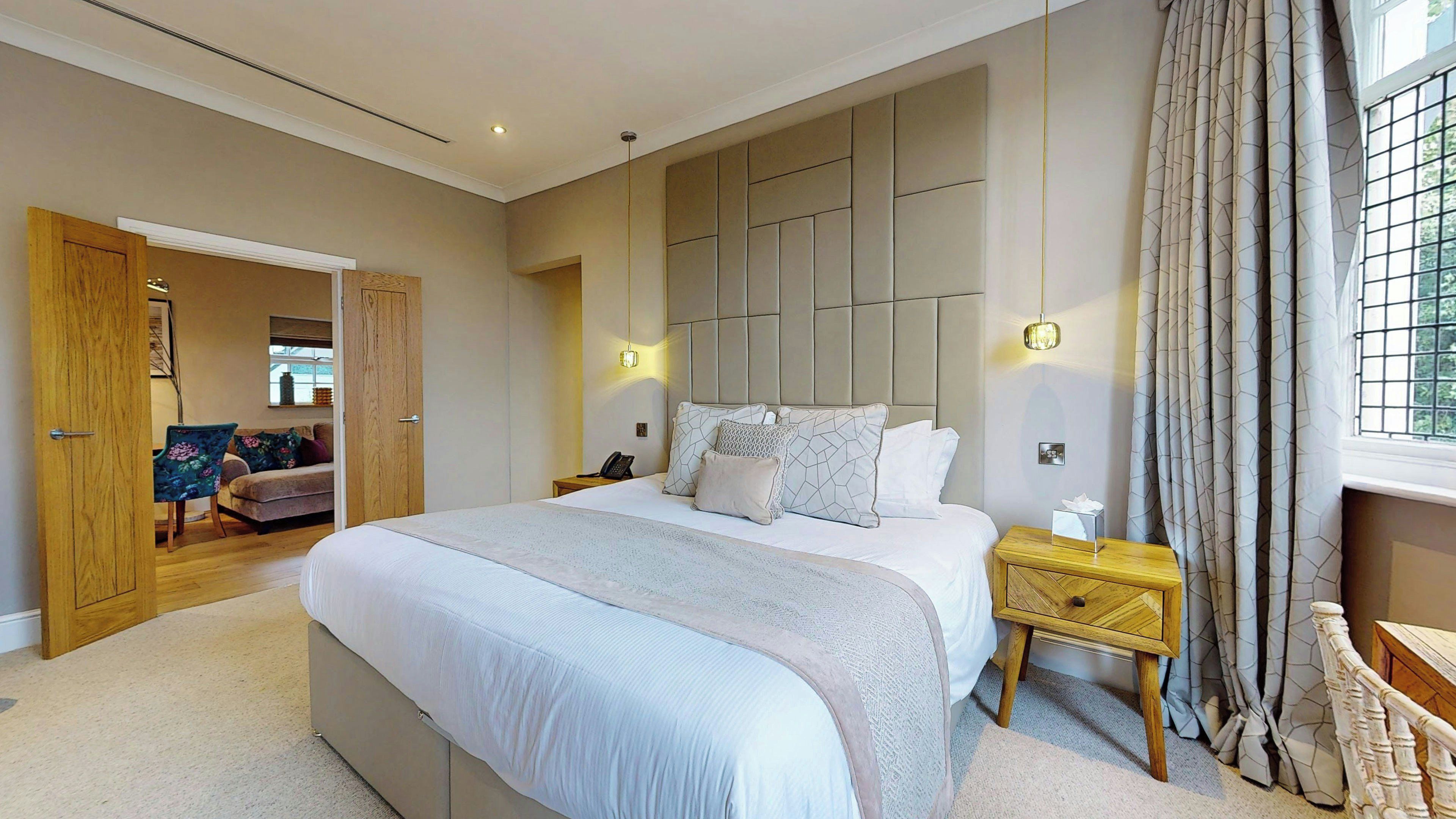A bedroom with a large bed, a wooden nightstand, a bedside table, a wardrobe, and a door leading to another room.