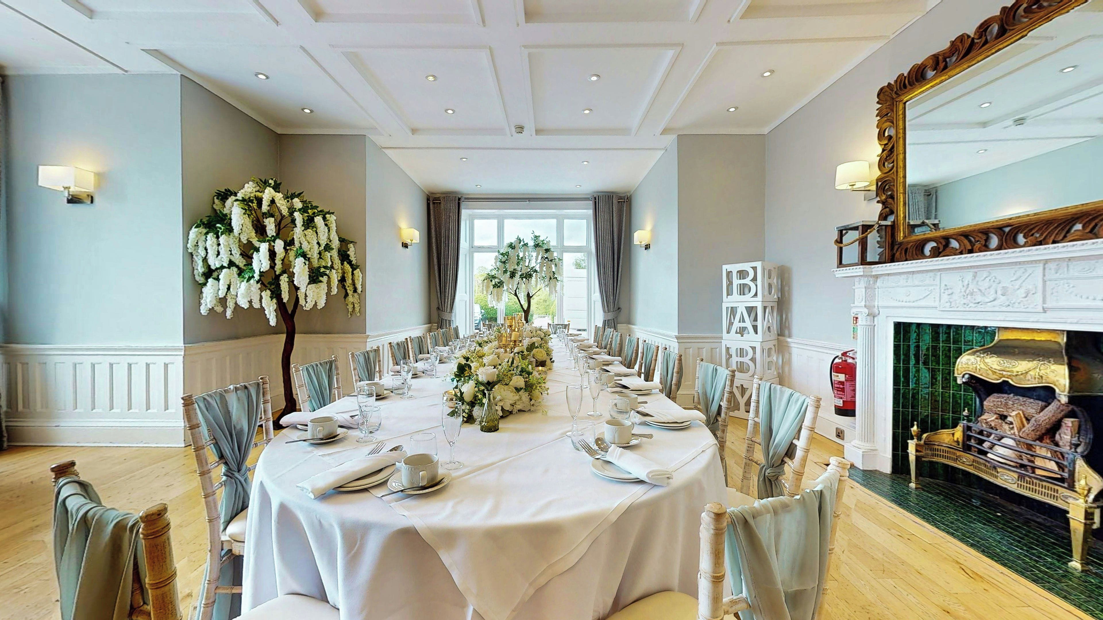 A formal dining room with a long table set for a meal, flanked by chairs with light blue drapes, a decorative white floral arrangement, and a fireplace with a mirror above it.