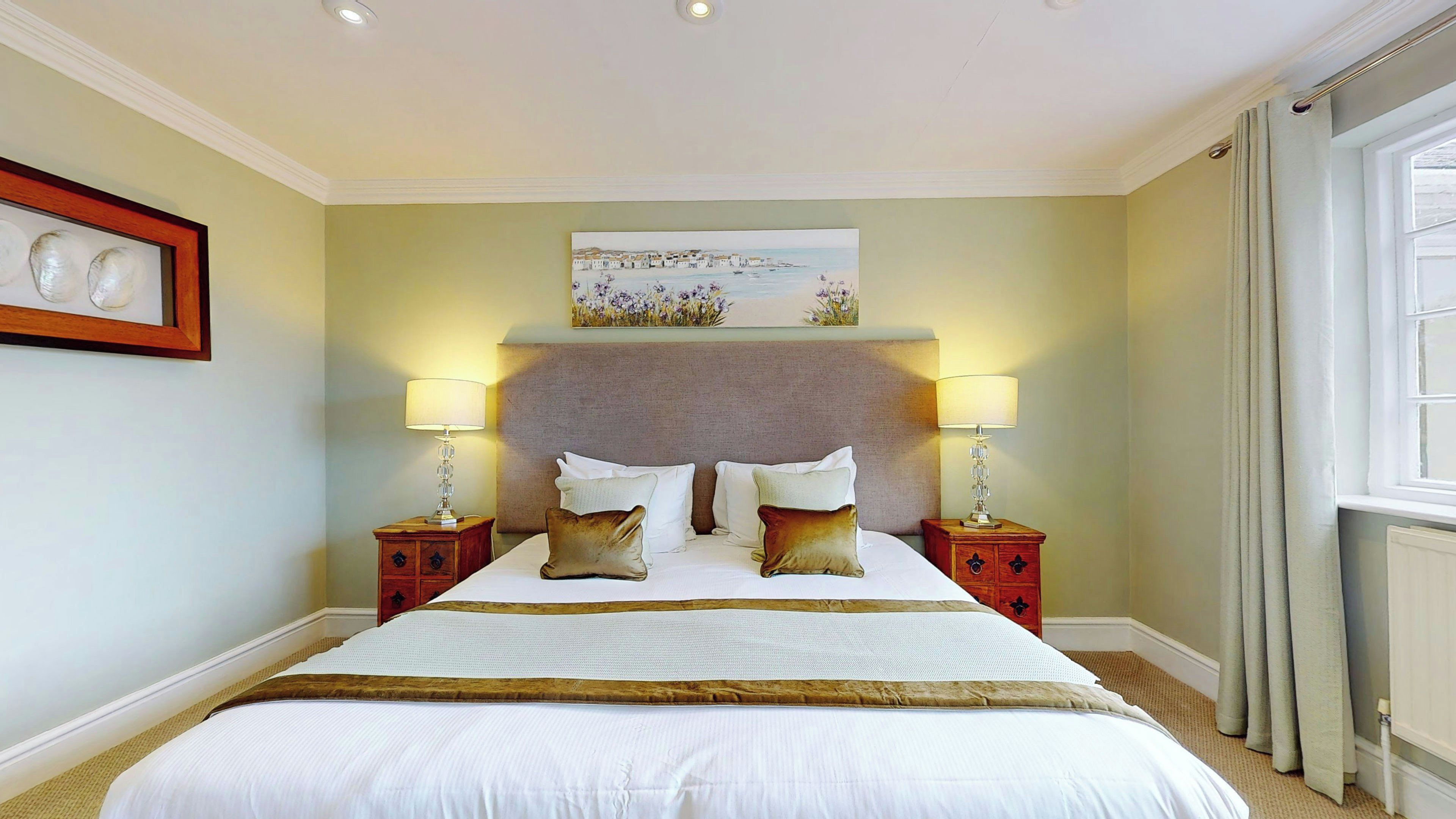 A hotel room with a large bed, two bedside tables with lamps, a painting above the bed, and a window with curtains.