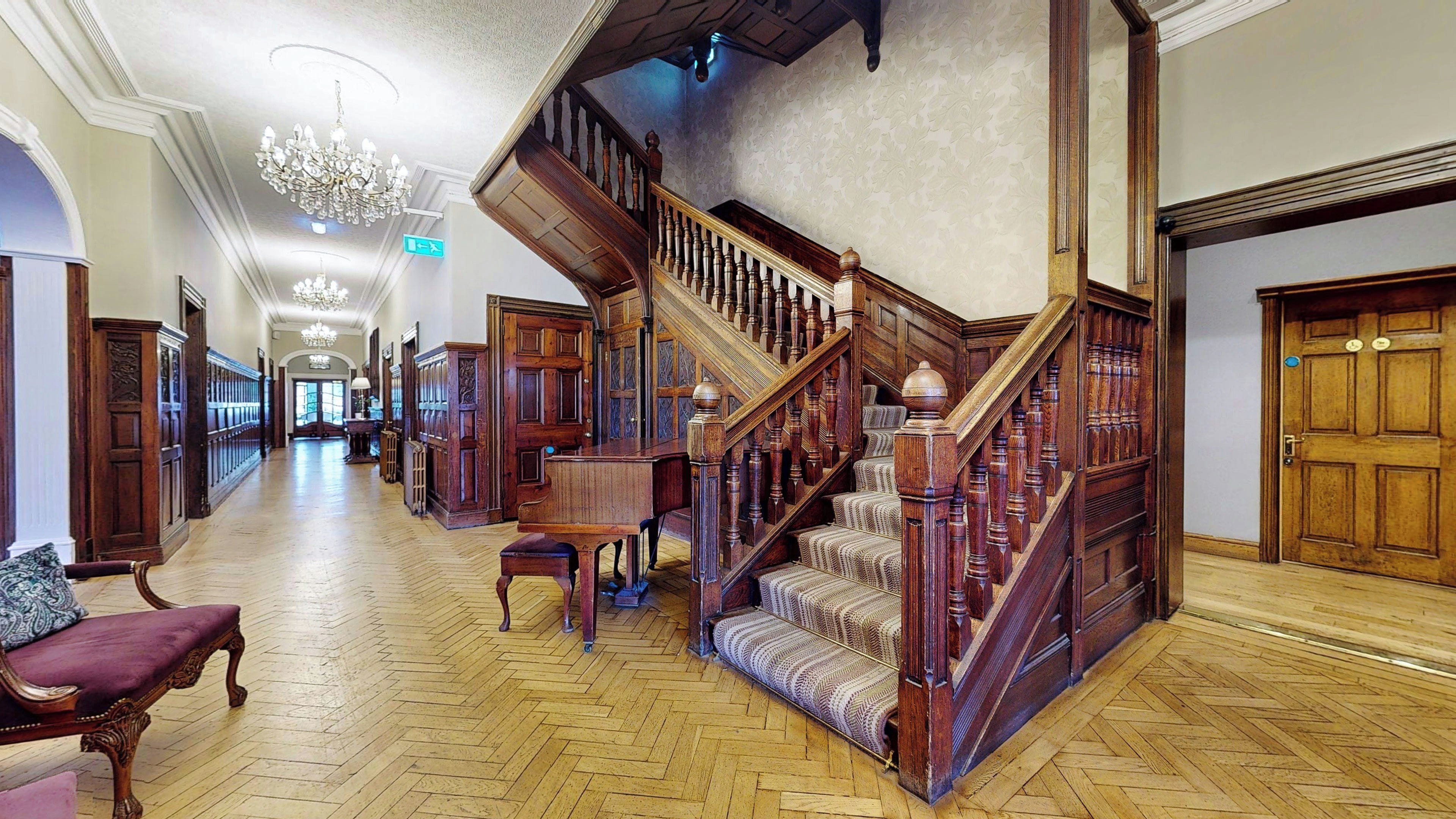 Un amplio pasillo con una escalera de madera, un candelabro y un banco.