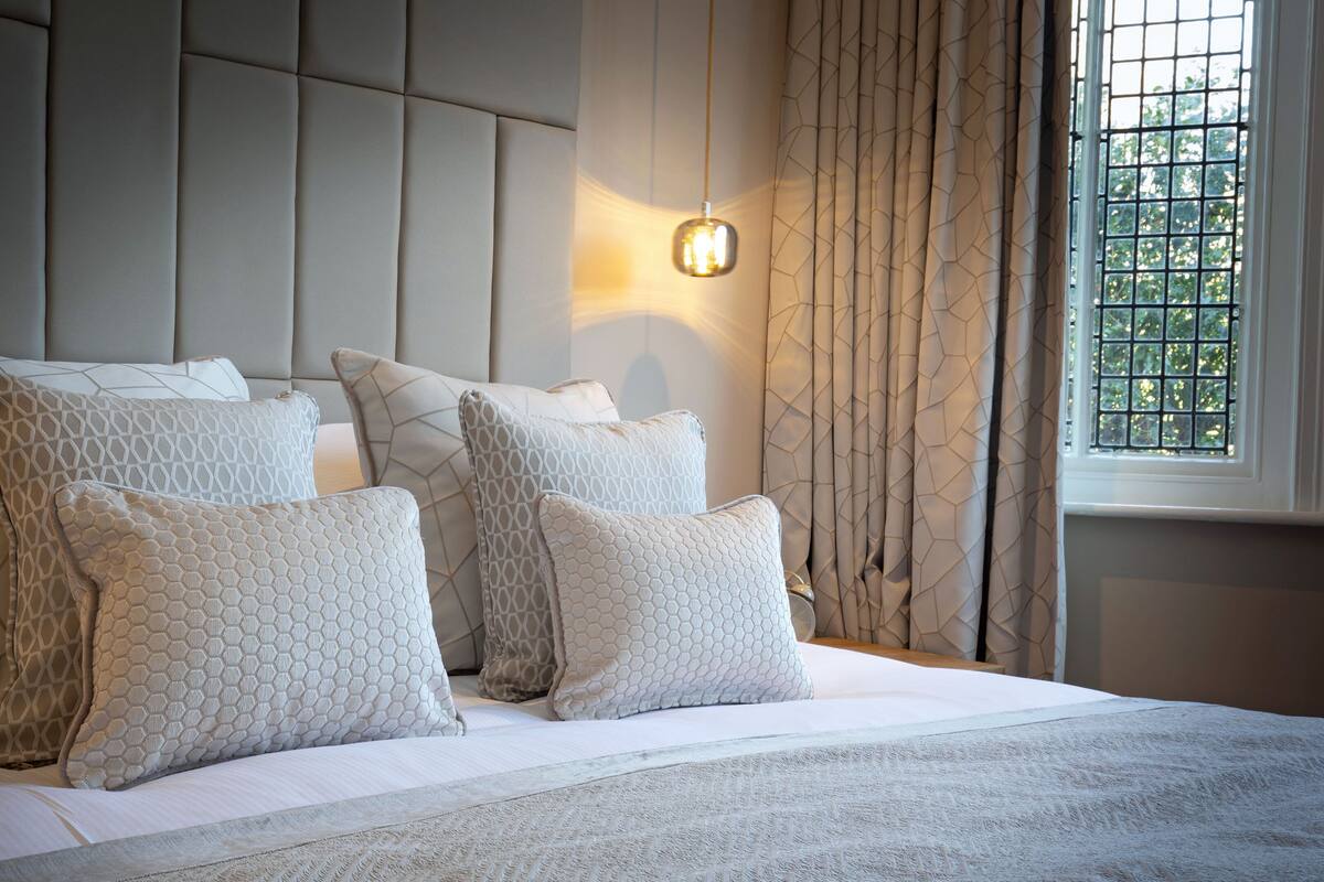 A neatly made bed with multiple pillows and a headboard, a bedside lamp, and a window with curtains.