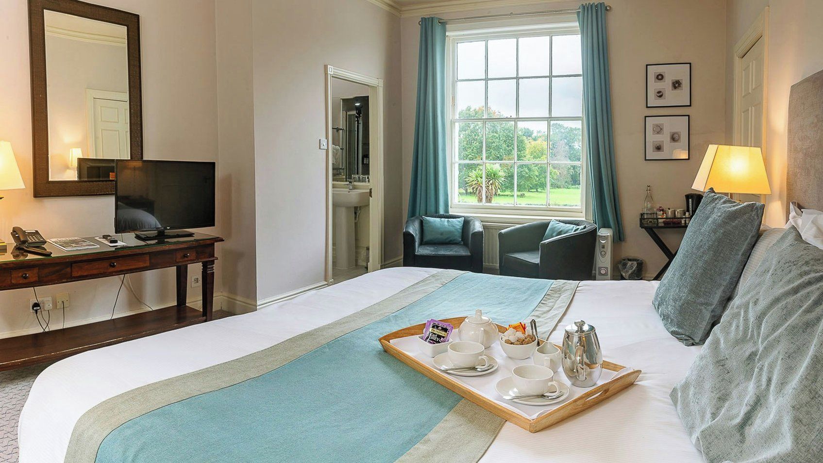 A hotel room with a bed, a desk with a computer, a chair, a window with curtains, and a tray with breakfast items.