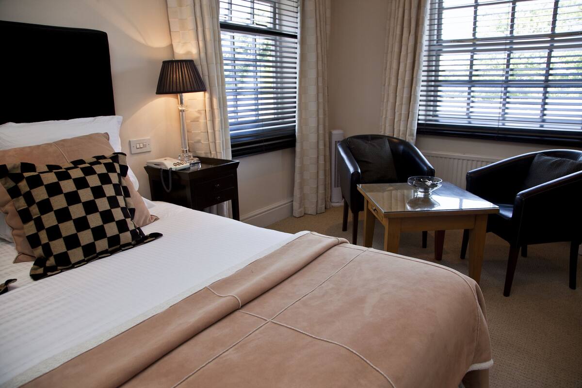 A neatly made bed with a checkered pillow, a bedside lamp, a table with a glass, and a chair in a hotel room with windows and curtains.