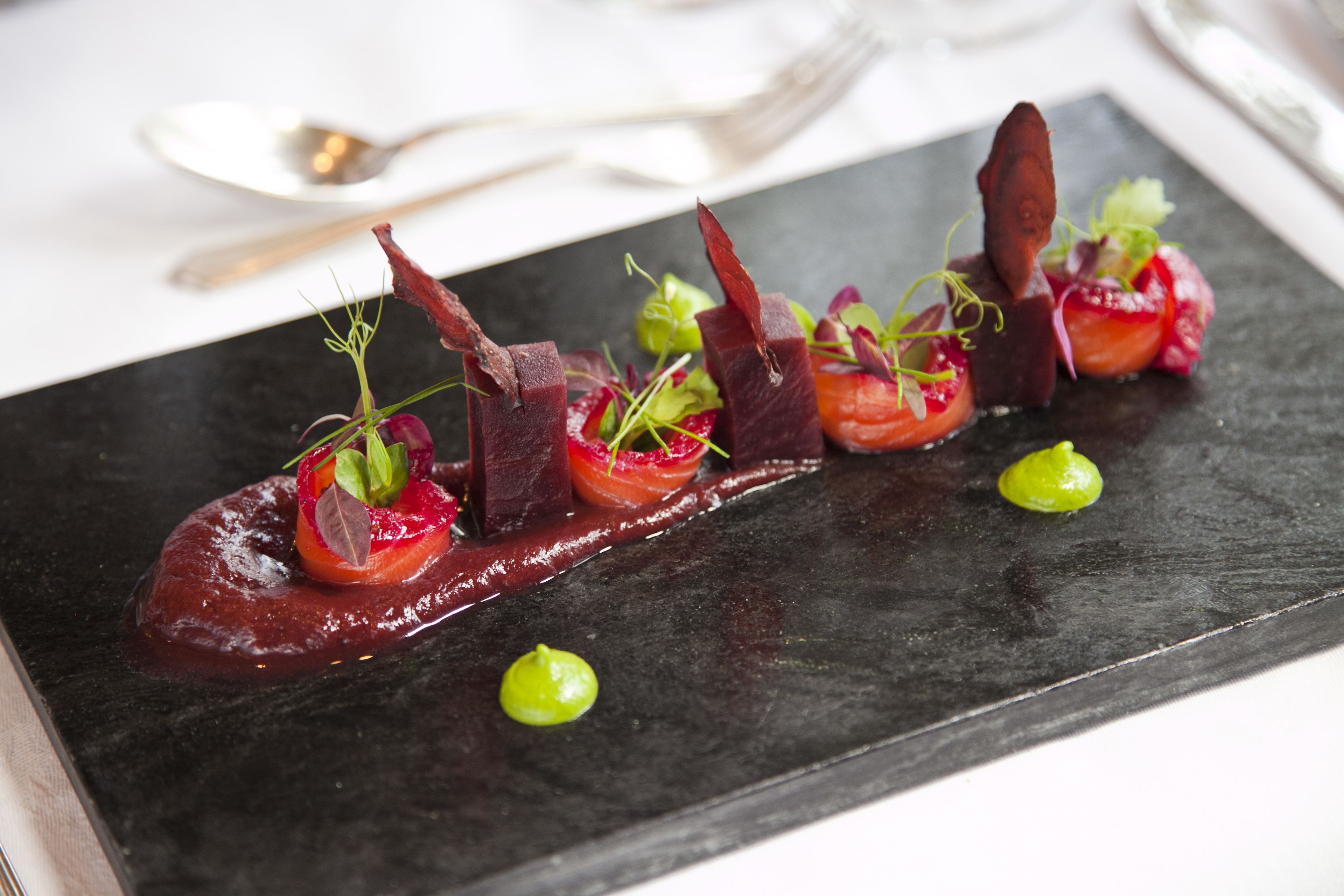 A black slate serving board with a dish of food, a spoon, and a fork on a white tablecloth.