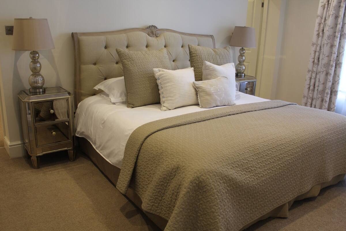 A neatly made bed with a beige quilt, two bedside tables with lamps, and a window with curtains.