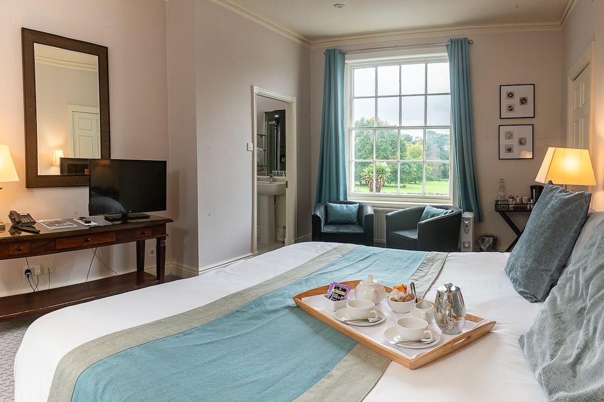 A hotel room with a large bed, a desk with a computer, a chair, a window with curtains, and a tray with breakfast items.