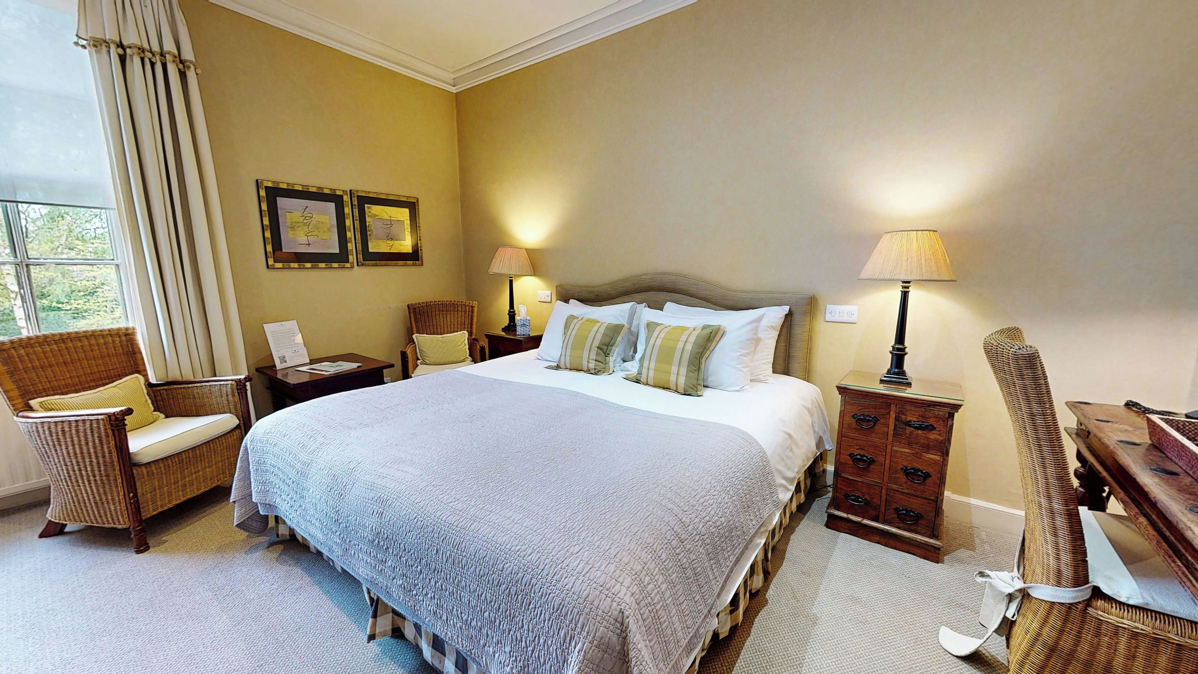 Una habitación de hotel con una cama grande, dos mesitas de noche con lámparas, una silla de mimbre, una mesita de madera y una pared con cuadros enmarcados.