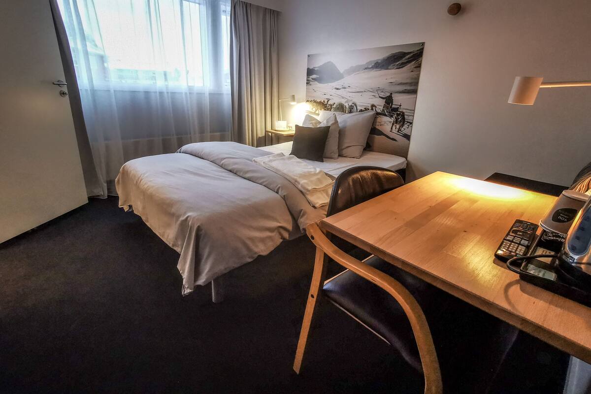 A hotel room with a bed, a desk, a chair, a nightstand, and a window with curtains.