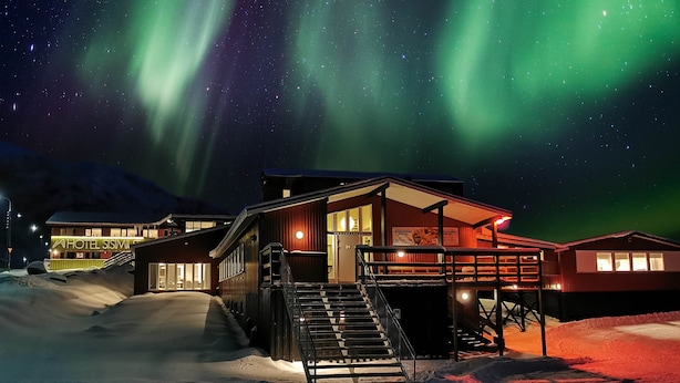 A hotel with a snow-covered ground, a staircase leading to the entrance, and a view of the night sky with auroras. - Hotel Sisimiut (Sisimiut)