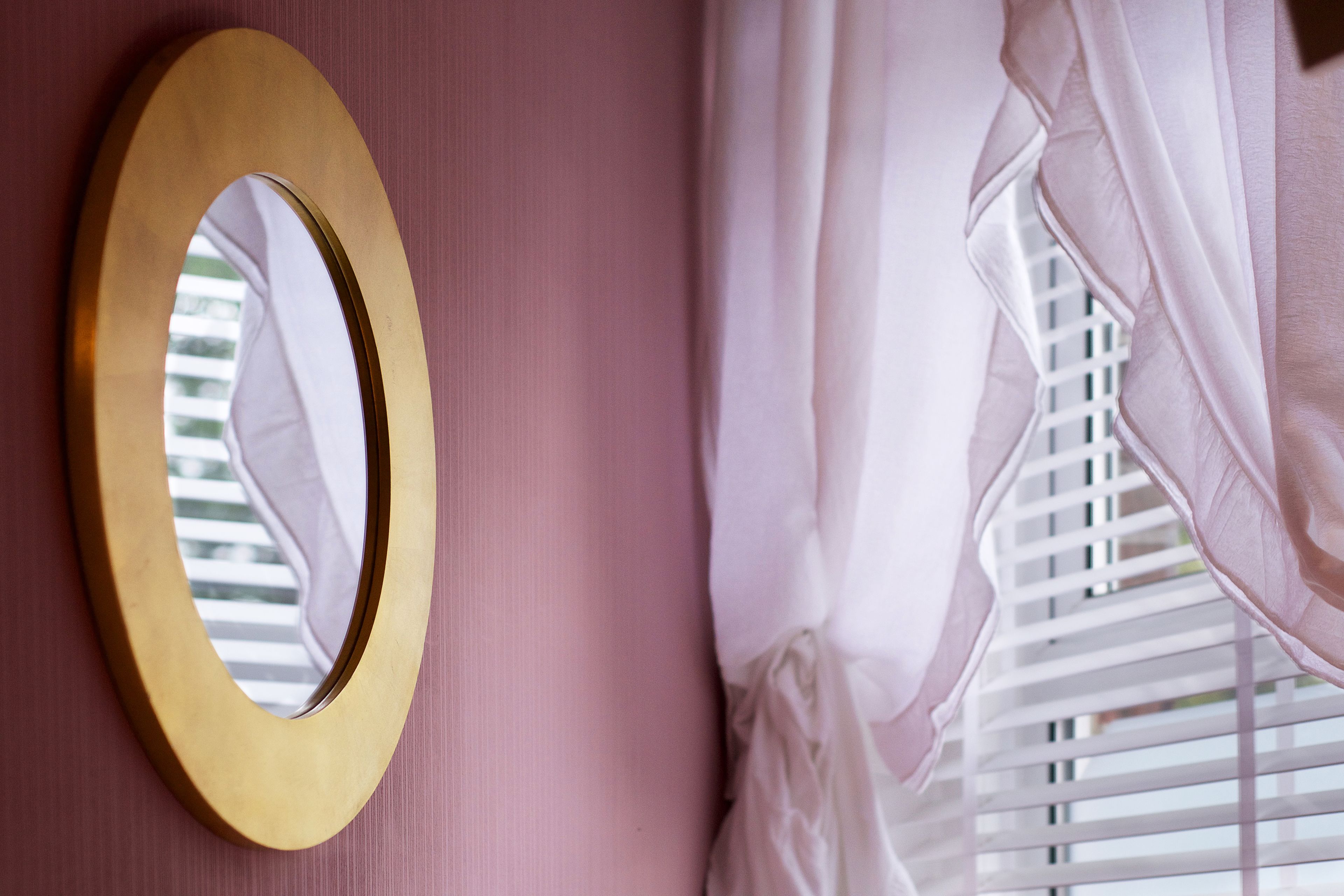 A room with a gold-framed mirror on a pink wall, white curtains, and a window with blinds.