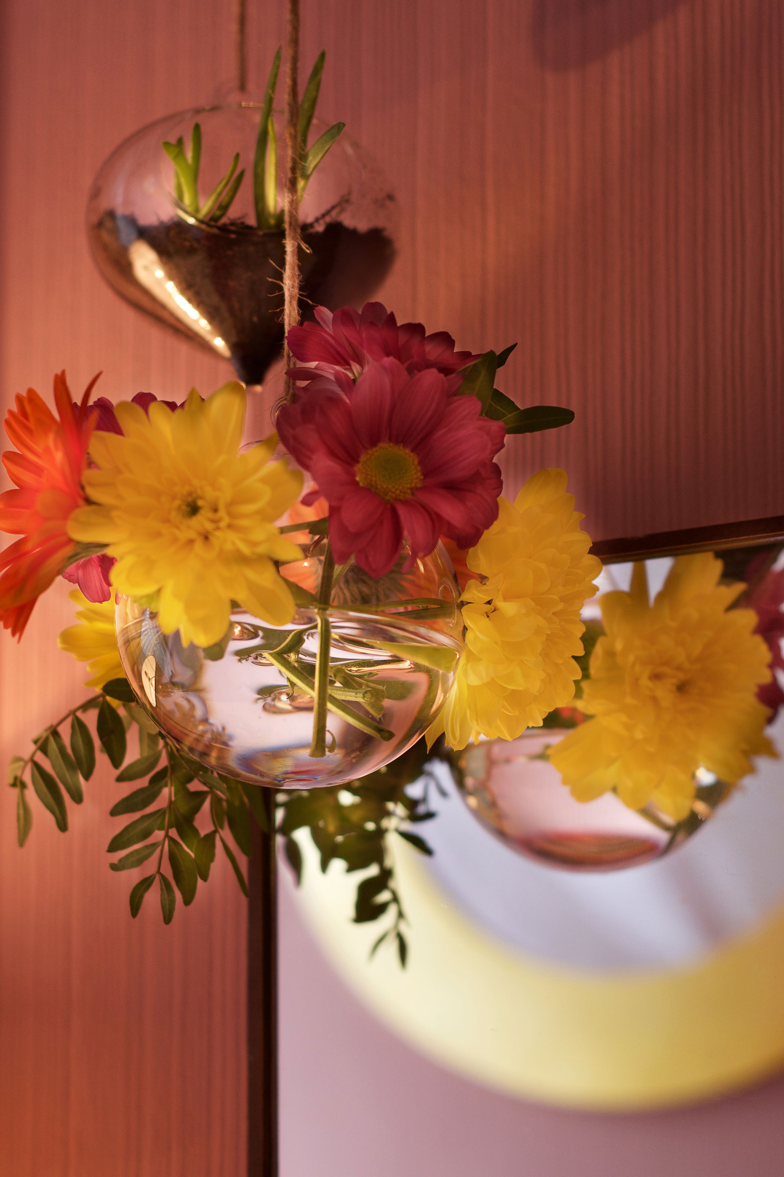 A clear glass vase with yellow and pink flowers, placed on a reflective surface.