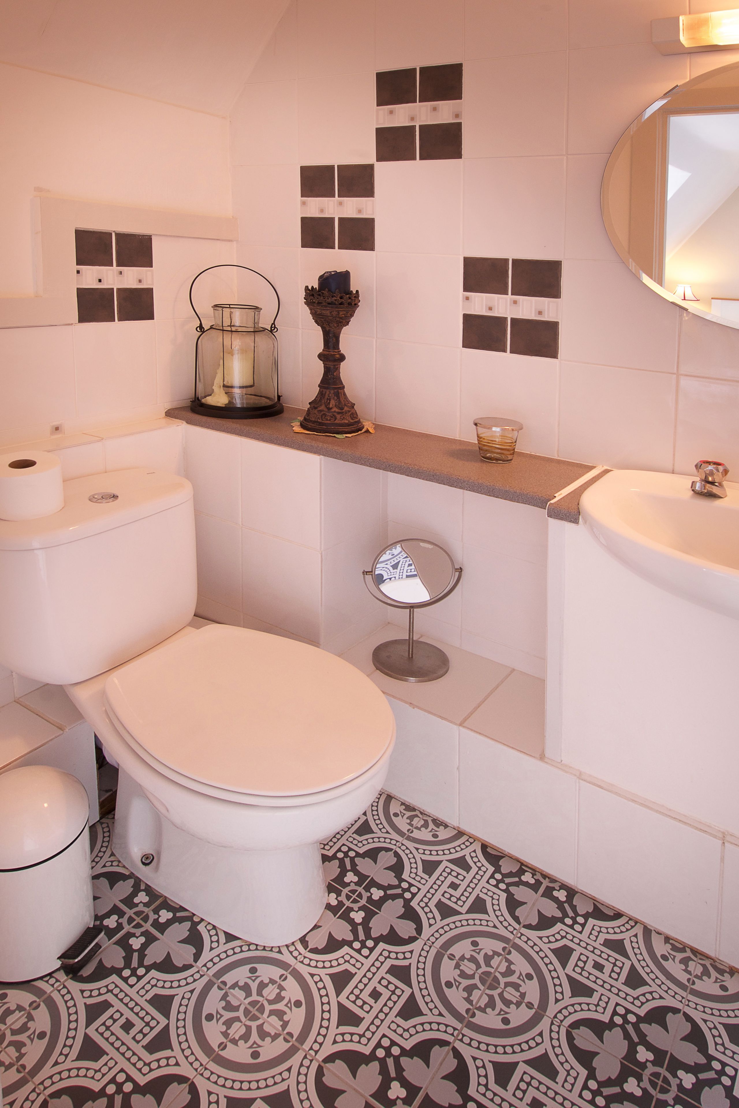 Une salle de bain avec des toilettes, un lavabo et un miroir. Le sol est recouvert d’un motif décoratif et une étagère contient une bougie et un petit bol.