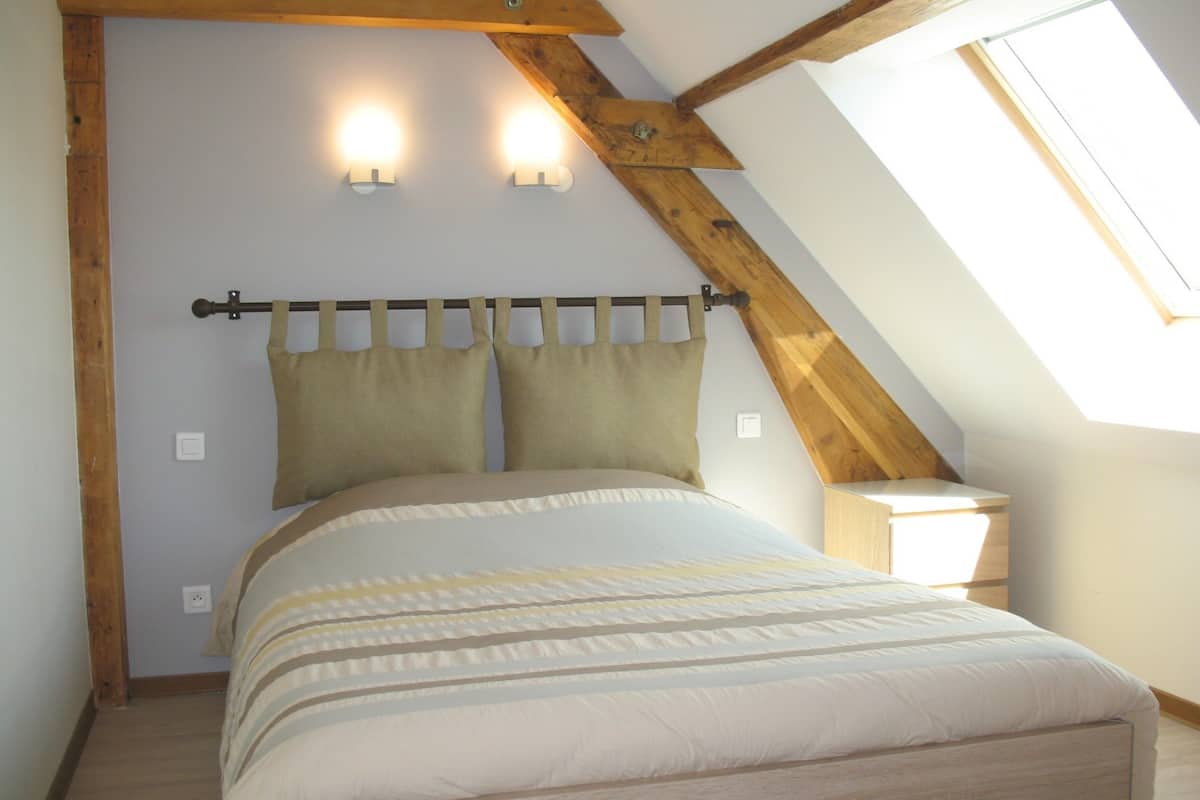 Ein Schlafzimmer mit schrägen Wänden, einem Bett mit gestreifter Bettdecke, zwei Wandleuchten und einem Fenster.